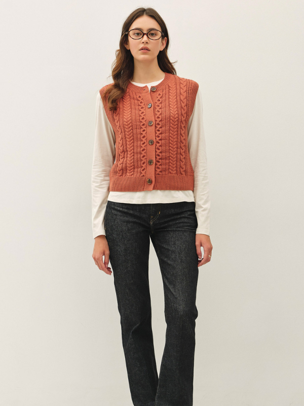 A woman modeling a rust-orange cable-knit button-front vest layered over a white long-sleeve shirt and dark gray jeans, standing against a plain light gray studio background.