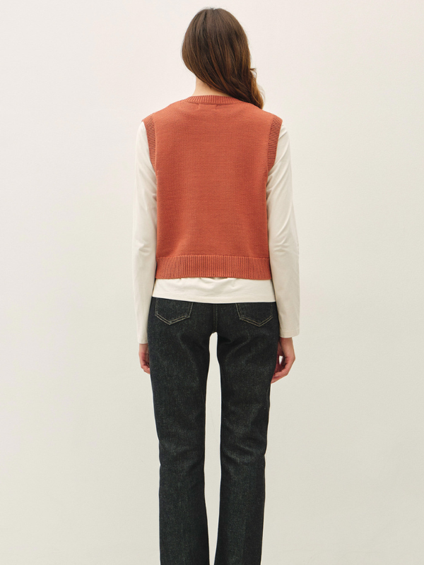 A woman viewed from behind wearing a rust-orange knit sweater vest over a cream long-sleeve shirt and dark charcoal jeans, set against a plain off-white studio background.