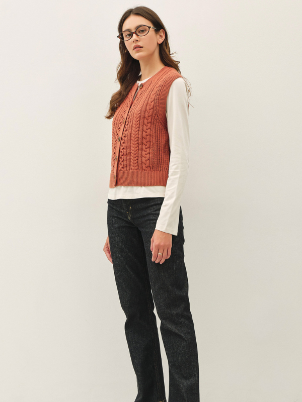A woman modeling a sleeveless rust-orange cable-knit vest over a white long-sleeve shirt and black jeans, standing against a plain light gray studio background.