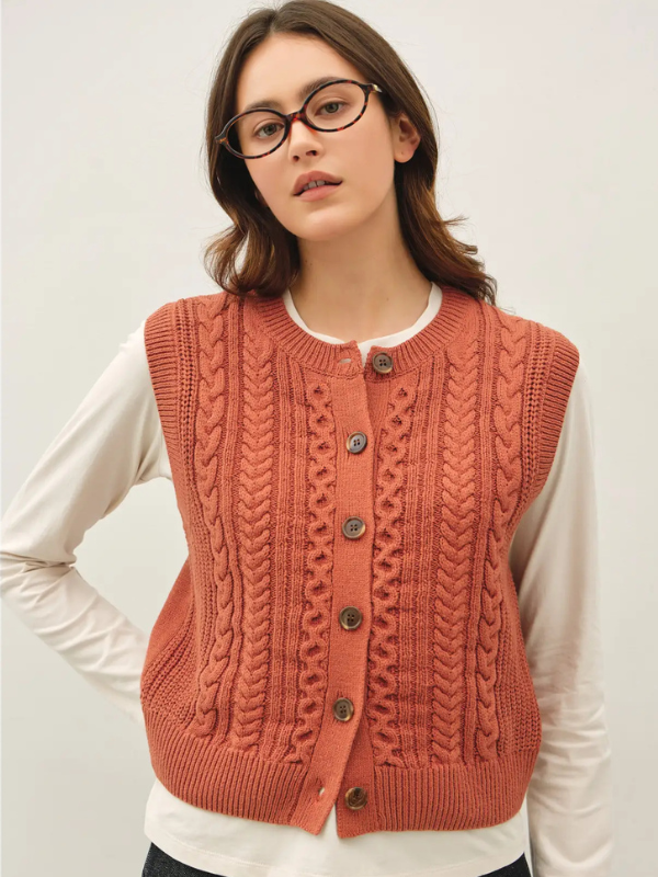 A woman wearing a rust-orange cable-knit cardigan vest over a white long-sleeve shirt, photographed against a plain light background.