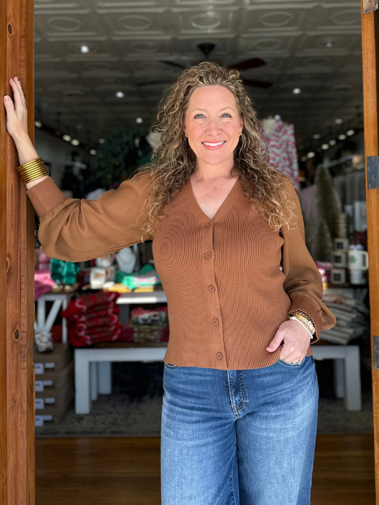 Woman standing in a store wearing a brown sweater and blue jeans, smiling.