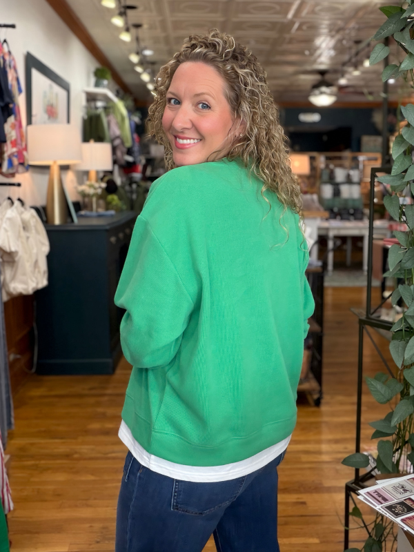 A woman with curly blonde hair wearing a bright green crewneck sweatshirt and dark blue jeans, photographed from behind inside a retail store.