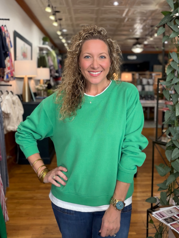 A smiling woman with curly blonde hair stands in a retail store wearing a bright green crewneck sweatshirt over a white t-shirt and blue jeans.
