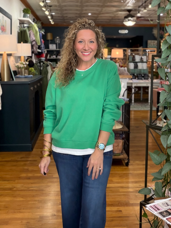 A smiling woman with curly hair stands in a retail store wearing a bright green crewneck sweatshirt over a white undershirt and dark blue jeans.