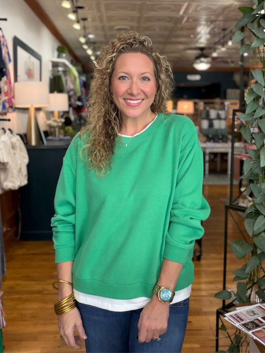 A smiling woman with curly blonde hair wearing a bright green crewneck sweatshirt over a white t-shirt and blue jeans, standing inside a retail store.