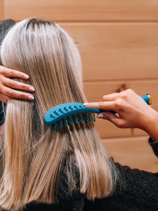 A teal blue hairbrush being used on a person's light blonde hair, shown from behind. The person is wearing a black knit sweater, and the background is a warm-toned wooden wall.