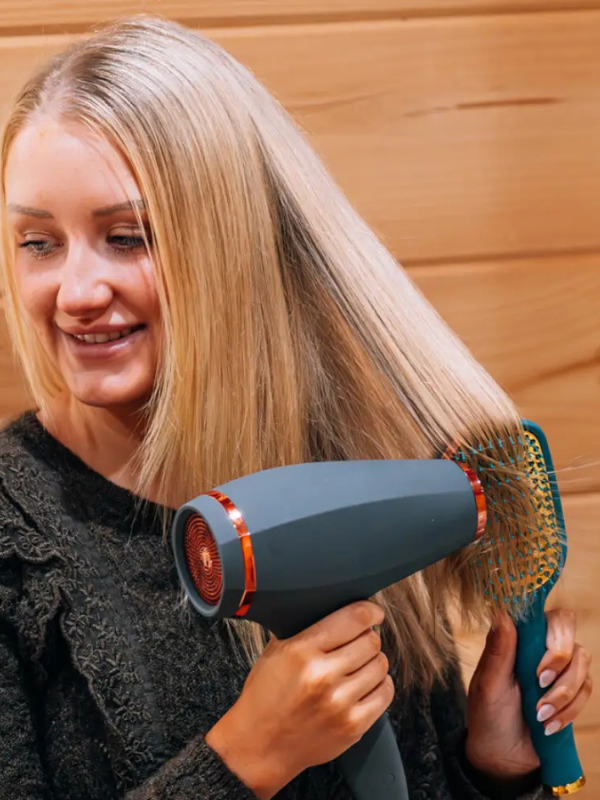A woman with long blonde hair uses a gray hair dryer while brushing her hair with a teal and gold paddle brush, set against a warm wood-paneled background.