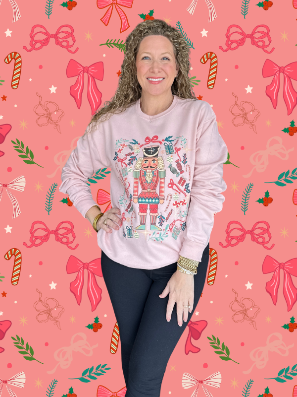 Woman wearing a pink sweatshirt with a nutcracker design on a pink background with Christmas decorations.