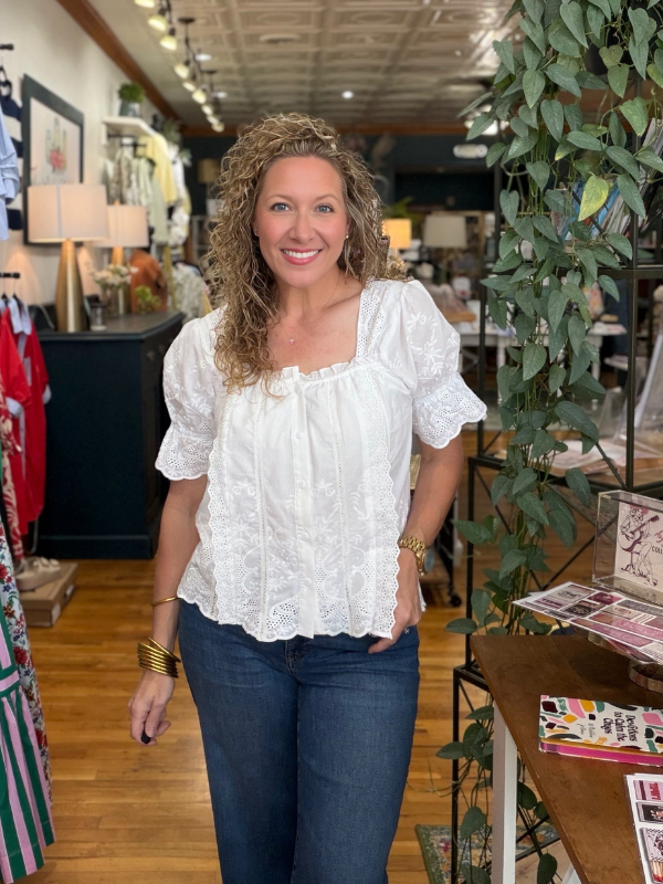 Woman in a white blouse and blue jeans standing in a store with plants and shelves in the background.
