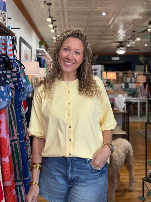 Woman in a yellow blouse and blue jeans standing in a store with clothing racks and a dog.