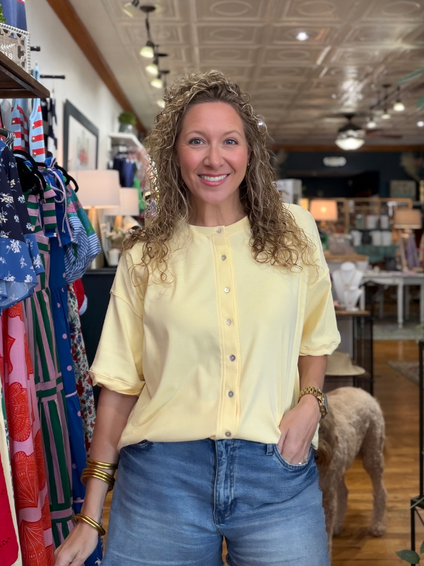 Woman in a yellow shirt and blue jeans standing in a store with colorful clothing around her.